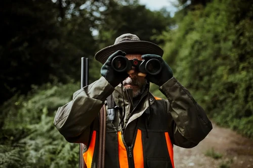 A man stands looking through his binoculars while hunting during the fall.