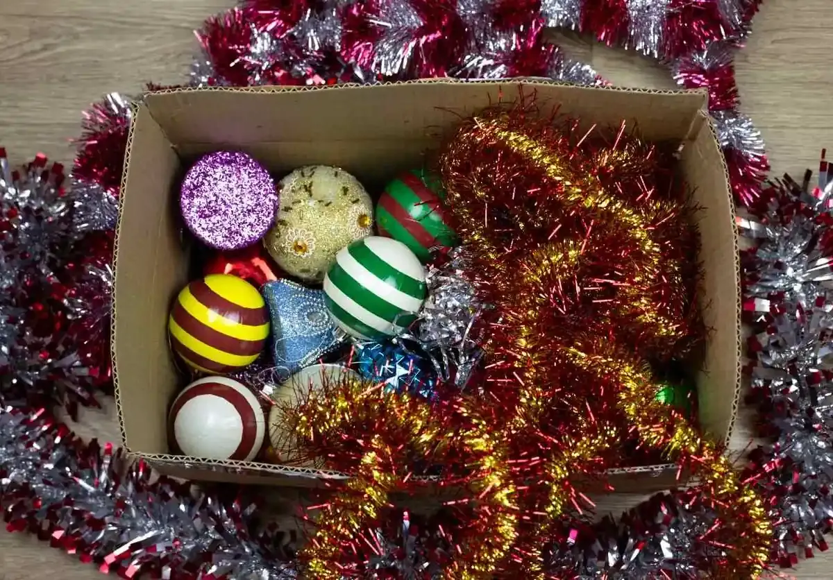 An open cardboard box filled with colorful Christmas ornaments and tinsel, showing mixed holiday decor stored loosely.